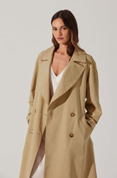 A woman wearing a long beige trench coat with wide lapels, double-breasted buttons, and side pockets. Underneath, she has on a white V-neck top. The coat has a classic, tailored style, and she has her hands in the coat’s pockets.